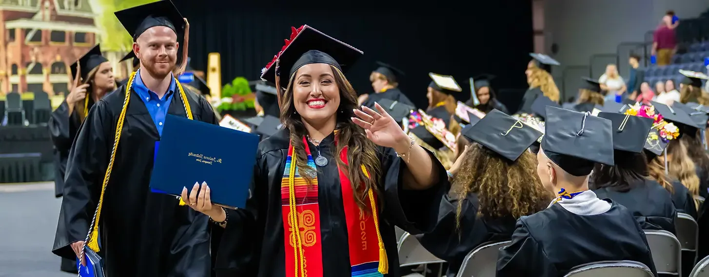 A woman in a graduation gown and cap smiling and waving during a graduation ceremony.